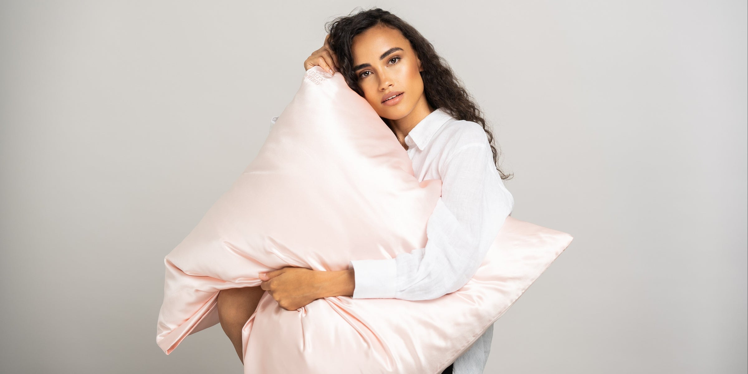 Schöne Frau mit dunklen Locken schmiegt ihr Gesicht an ein rosafarbenes Seidenkissen von CURLY N COVERED