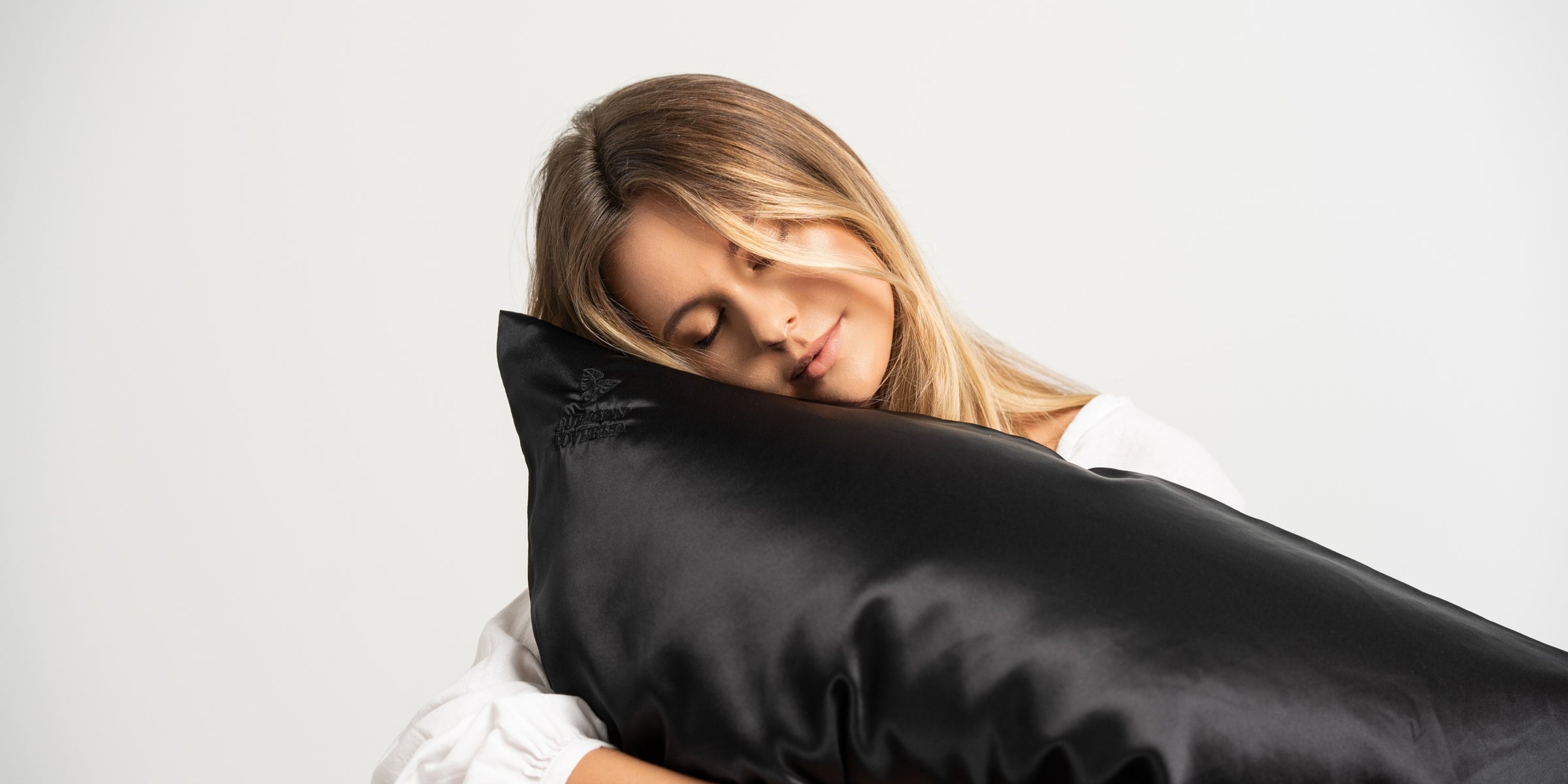 Schöne blonde Frau schmiegt sich an ein Kissen mit einem schwarzen Seidenkissenbezug aus reiner Maulbeerseide von CURLY N COVERED