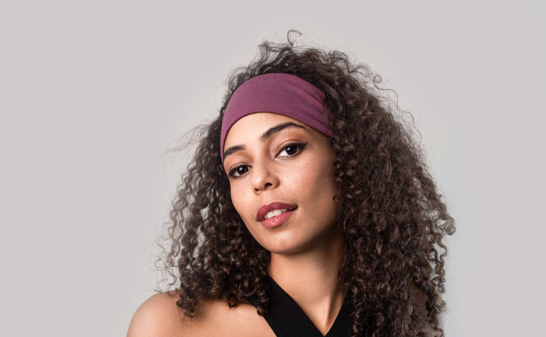 Eine junge Frau mit offener dunkler Lockenpracht trägt ein violettes Stirnband aus Bambus von Curly'N'Covered und schaut lässig in die Kamera