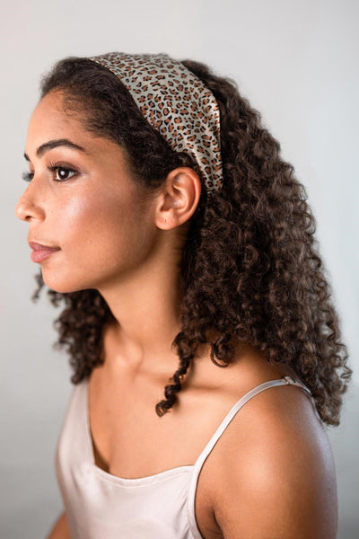 Wunderschöne Frau mit dunklen Locken trägt ein elegantes breites Haarband mit Leo Optik aus 100% Seiden-Satin von Curly N Covered als Stirnband
