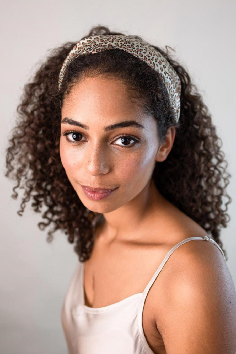 Schöne Frau mit dunklen Locken trägt ein elegantes breites Haarband mit Leo Optik aus 100% Seiden-Satin von Curly N Covered als Stirnband