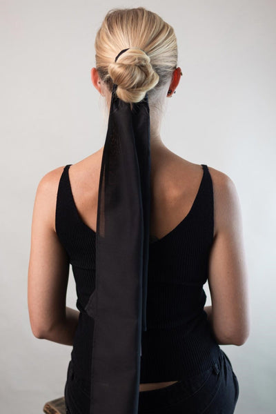 Frau mit langen blonden Haaren hat sich mit einem schwarzen Hair Scarf aus Baumwoll-Seide von Curly N Covered einen Dutt gebunden und der Haar-Schal fällt vom Kopf leicht über ihren Rücken