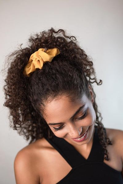 Lockerer Zopf mit dunklen lockigen Haaren, zusammengehalten von einem Scrunchie aus Baumwollseide von Curly'N'Covered in der Farbe Gold